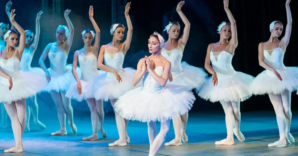 Bailarinas em tutus brancos se apresentam no palco, com uma dançarina no centro da imagem. Eles estão graciosamente posicionados com os braços levantados, colocados contra um fundo mal iluminado, sugerindo um cenário de teatro. A iluminação projeta sombras suaves, realçando a elegância da cena.