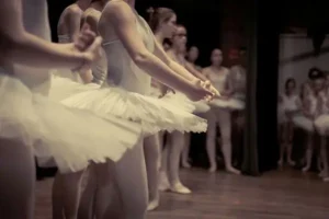 Bailarinos em tutus brancos estão em uma fila em um palco de madeira mal iluminado, se preparando para uma apresentação. Outros dançarinos são visíveis ao fundo.