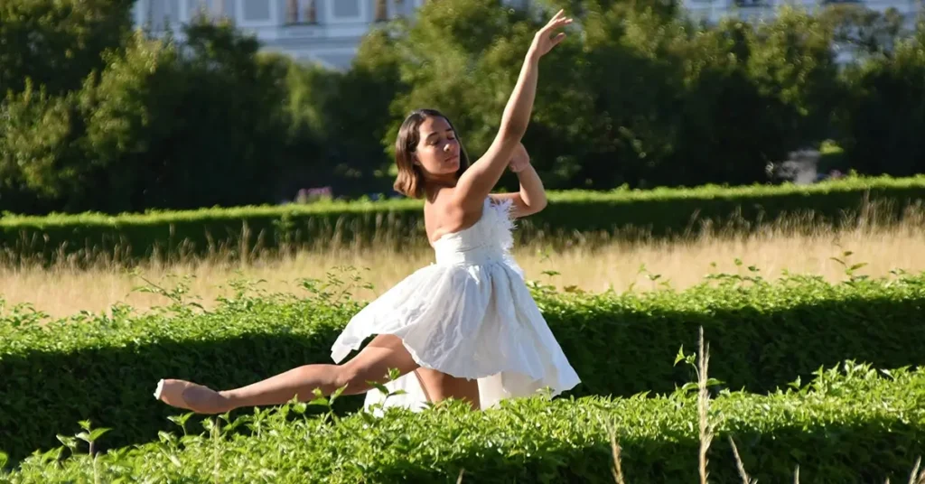 Uma dançarina de vestido branco posa graciosamente com uma perna estendida, cercada por sebes verdes e grama.