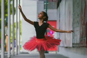 Uma jovem bailarina em um tutu vermelho vibrante e top preto se apresenta na ponta dos pés com um braço graciosamente levantado. Ela está posicionada em uma calçada da cidade sob uma passarela coberta moderna. Um mural decora a parede atrás dela, adicionando cor à cena.
