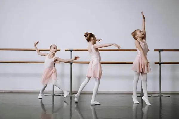 Três meninas em trajes de balé, sorrindo e posando em um estúdio de dança, cercadas por espelhos e barras de balé.