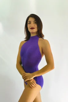 Bailarina a usar o Collant Frente Única Imaginário roxo da Lucia Caliman, com as mãos suavemente apoiadas na perna, a exibir a elegância da gola alta e a textura do tecido canelado.