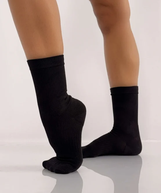 Bailarina usando Meia Contemporâneo Jazz preta da Lucia Caliman, em posição de meia-ponta alta, destacando o ajuste perfeito no arco do pé.