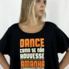 Jovem dançarina usando a Camiseta Cropped Arara preta da Lucia Caliman. A peça possui decote canoa e estampa frontal nas cores laranja e creme com a frase "Dance como se não houvesse amanhã" e detalhes de notas musicais. A modelo posa com as mãos na cintura, destacando o caimento leve da peça.