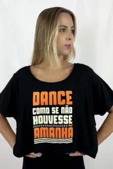 Jovem dançarina usando a Camiseta Cropped Arara preta da Lucia Caliman. A peça possui decote canoa e estampa frontal nas cores laranja e creme com a frase "Dance como se não houvesse amanhã" e detalhes de notas musicais. A modelo posa com as mãos na cintura, destacando o caimento leve da peça.