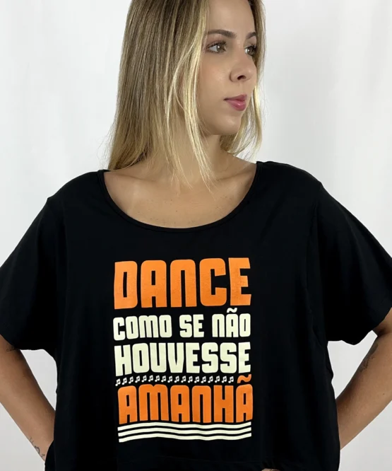 Jovem dançarina usando a Camiseta Cropped Arara preta da Lucia Caliman. A peça possui decote canoa e estampa frontal nas cores laranja e creme com a frase "Dance como se não houvesse amanhã" e detalhes de notas musicais. A modelo posa com as mãos na cintura, destacando o caimento leve da peça.