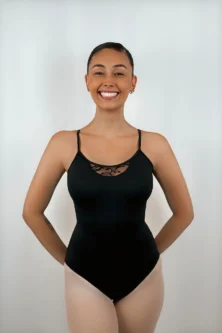 Bailarina sorridente usando o Collant Alça Trançada preto da Lucia Caliman, com as mãos nas costas, destacando o decote frontal que possui um delicado detalhe em renda floral.