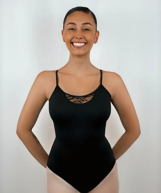 Bailarina sorridente usando o Collant Alça Trançada preto da Lucia Caliman, com as mãos nas costas, destacando o decote frontal que possui um delicado detalhe em renda floral.