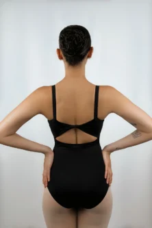 Bailarina de costas vestindo o Collant Costas Torcida preto da Lucia Caliman, exibindo o detalhe de tecido torcido no centro das costas nuas e as alças largas, em uma pose com as mãos na cintura.