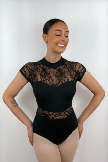 Bailarina sorrindo usando o Collant Manga Japonesa preto da Lucia Caliman. A imagem destaca a gola alta, as delicadas mangas curtas em renda floral, o decote ilusão em formato de coração no busto e a faixa de renda na cintura, com a modelo posando com as mãos nos quadris.