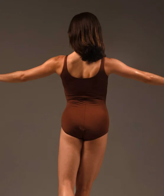 Bailarina de costas com os braços abertos vestindo o Collant Terra Áurea Essências da Terra marrom, exibindo o decote em U profundo e o caimento perfeito da peça.